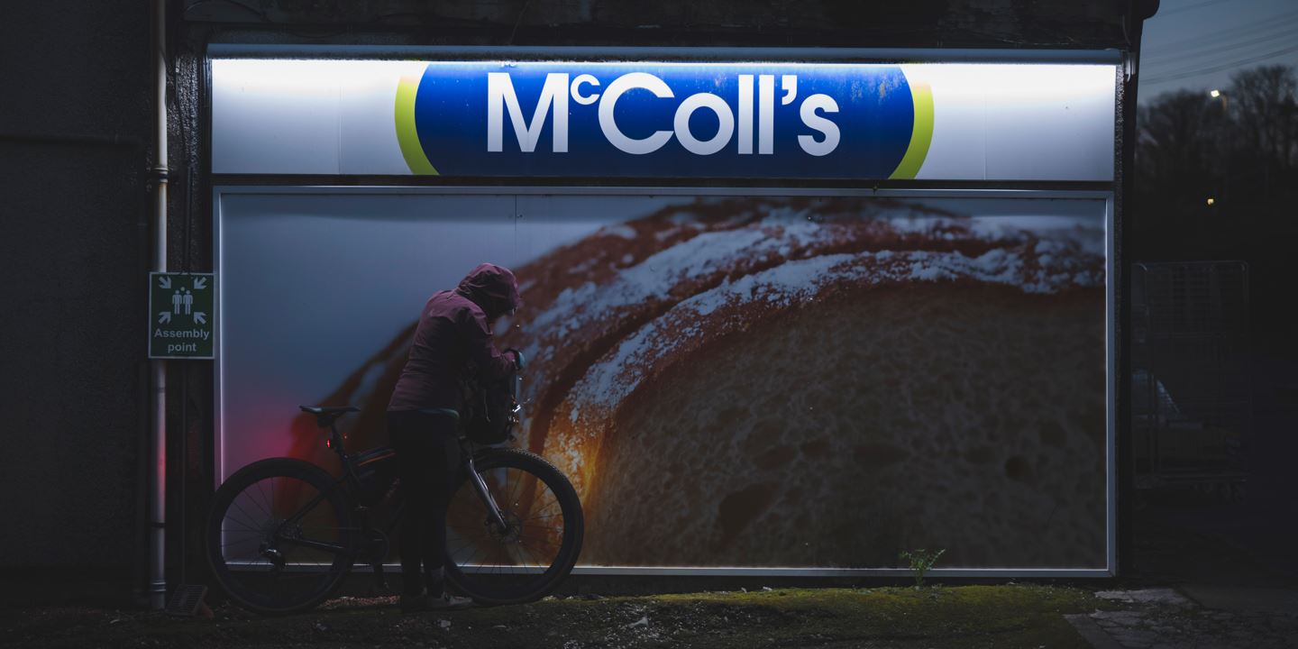 Rachael Walker leaning her bike against a McColl's neon-lit billboard with a picture of bread at night.